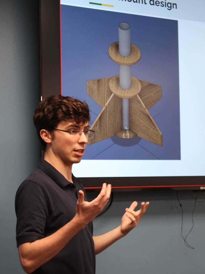 Presenting at a critical design review, mid-gesture, in front of a projected slide headed “mount design”. The slide shows a CAD render of a rocket motor mount: a motor tube running through two plywood centering rings, with the fin can bonded around them.