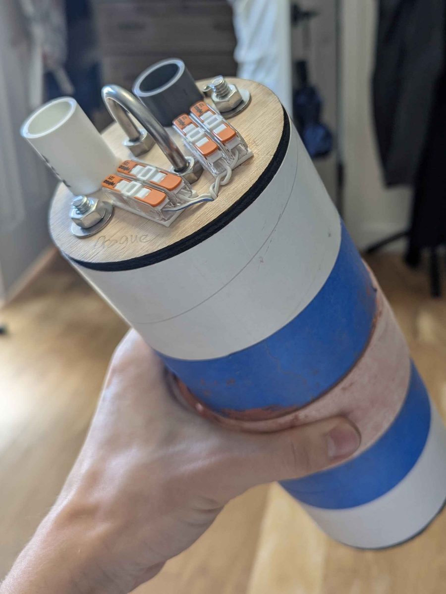 The avionics bay closed up in its coupler, held in one hand indoors before the launch. The plywood bulkhead on top carries two ejection charge wells, a steel U-bolt and four lever-nut terminals, all clean and unfired.