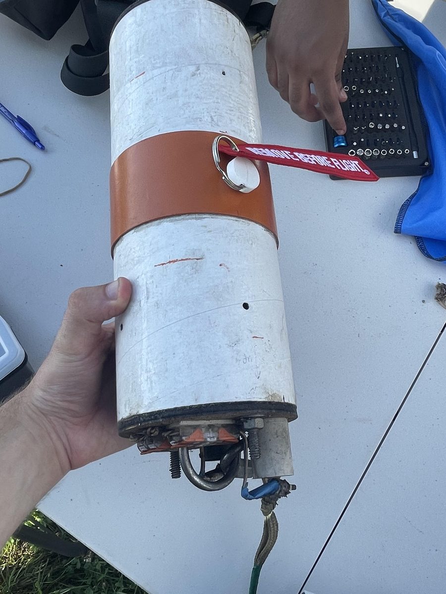 The same bay held over a table on the launch field after recovery. Its Remove Before Flight tag is still on the arming pin, the shock cord is still knotted to the U-bolt on the sooted bulkhead, and the threaded rods and nuts holding the stack together are all still in place.