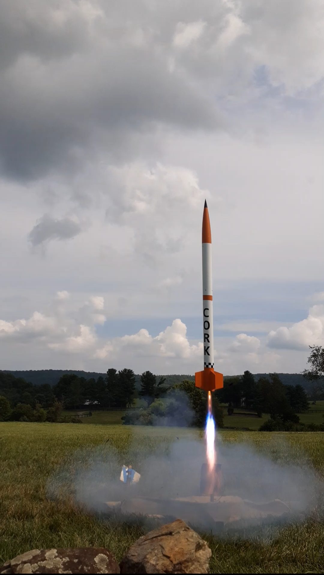 The CORK rocket climbing off its pad under thrust, exhaust plume beneath it and smoke spreading low across the grass.
