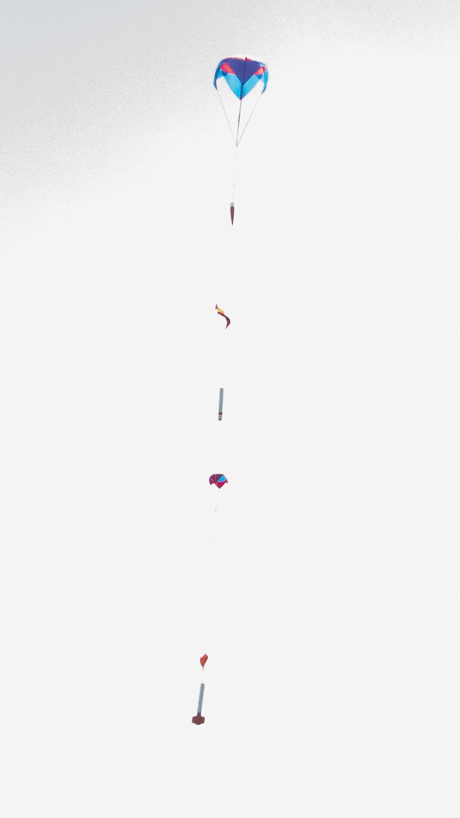 The vehicle descending against a white overcast sky: a large canopy at the top, the airframe hanging below it, a smaller canopy further down, and the lower airframe section beneath that — all on one vertical line.
